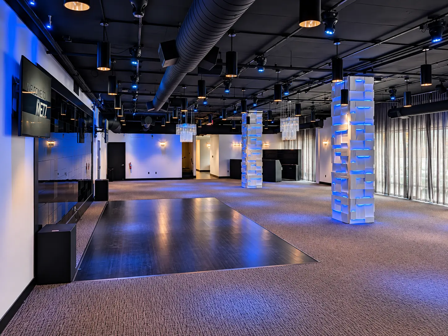 Wide corner view of the main event space at The MEZZ, with the dark wood dance floor, crystal chandeliers and blue-lit architectural columns beneath a black ceiling